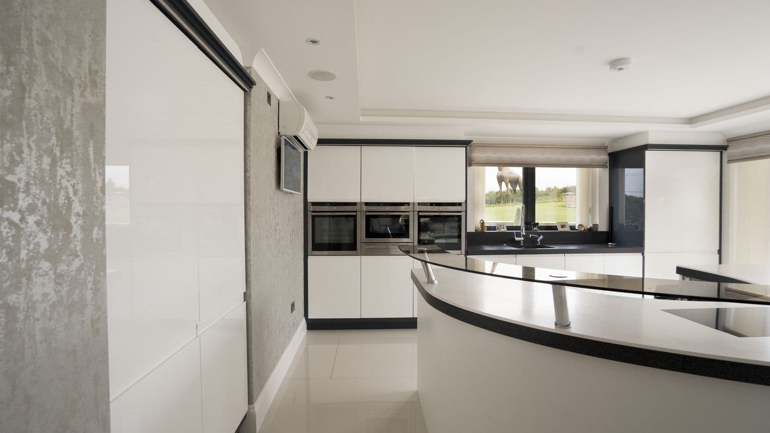 Straight on view of the gloss white Corian designer kitchen, notice the large doors sunk within the left wall housing the integrated fridge/freezer.