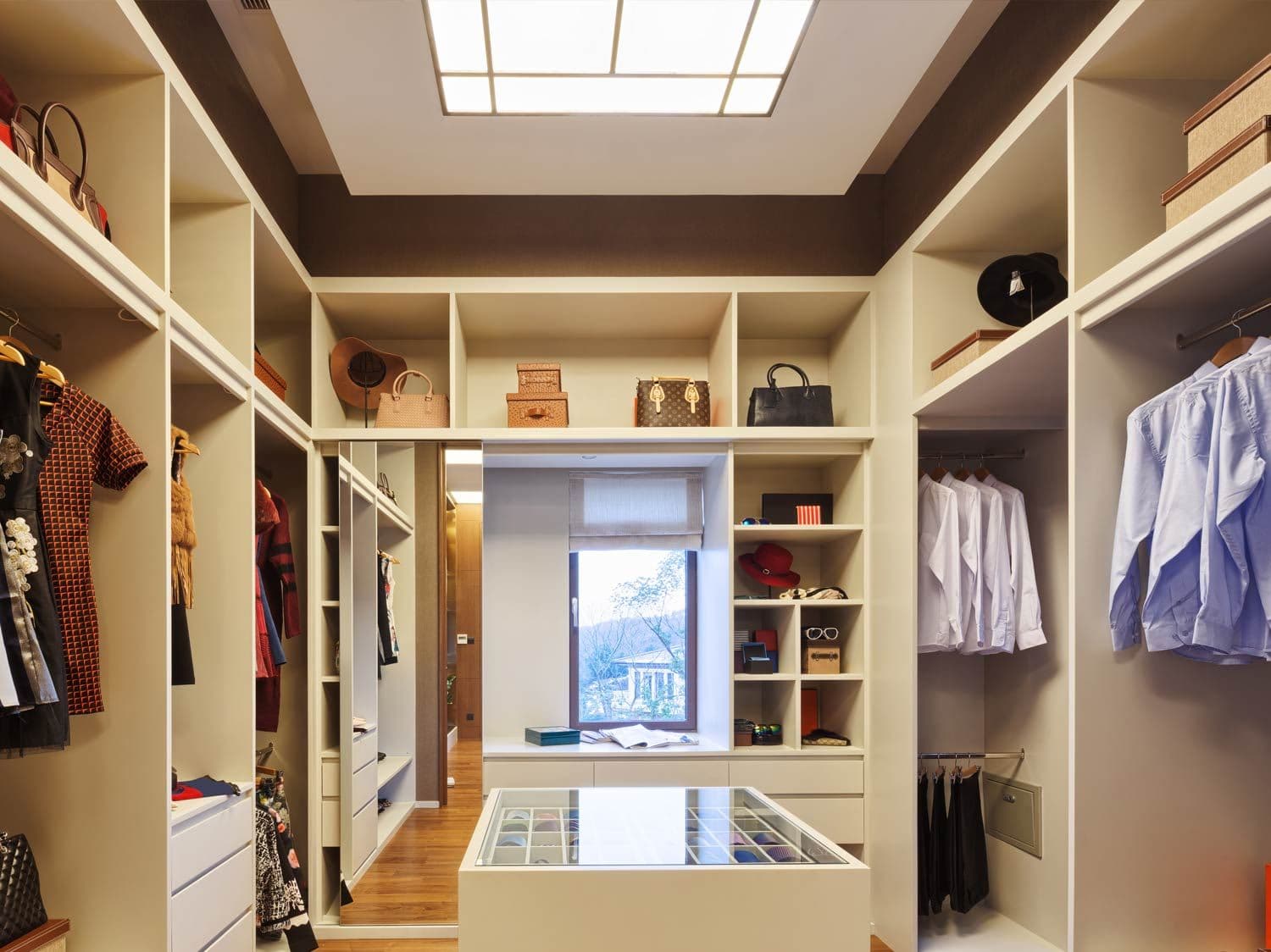 Large walk in wardrobe finished in white with sliding mirror door and a dressing table. In the centre of the room is a glass topped storage solution for housing ties and other accessories.